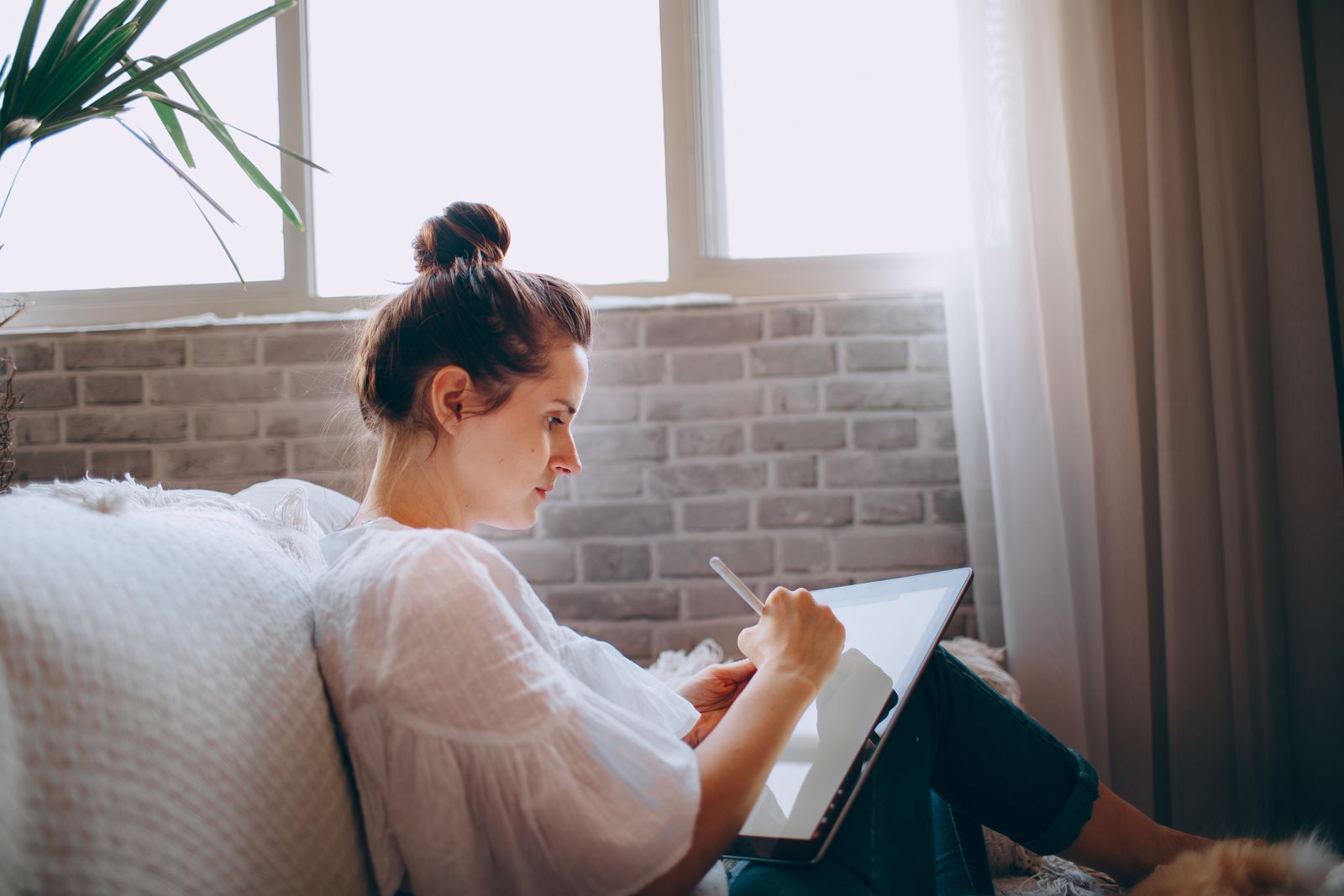 Woman sitting comfortably on a sofa using a digital tablet by a bright window.