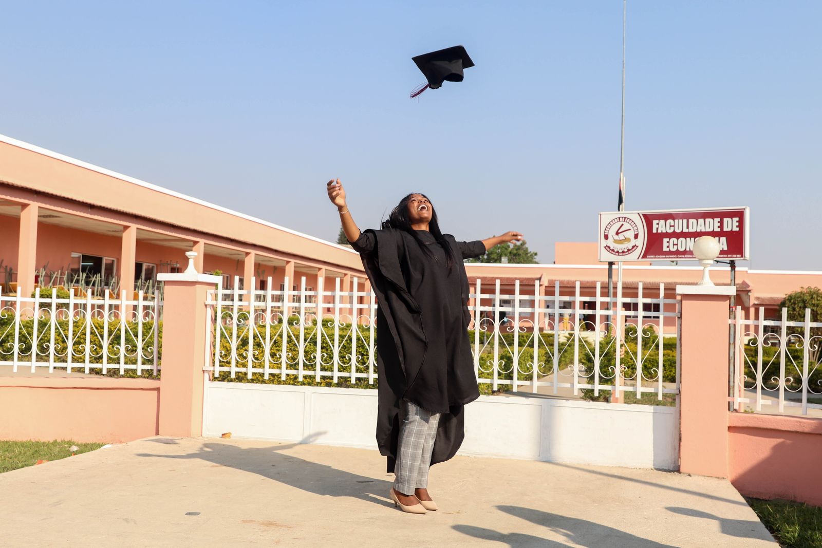 A jubilant graduate celebrating outside Huambo's university with a gown and mortarboard tossed in the air.