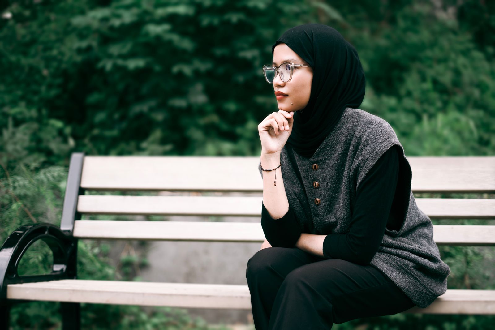 A woman in a hijab sits pensively on a park bench surrounded by greenery.