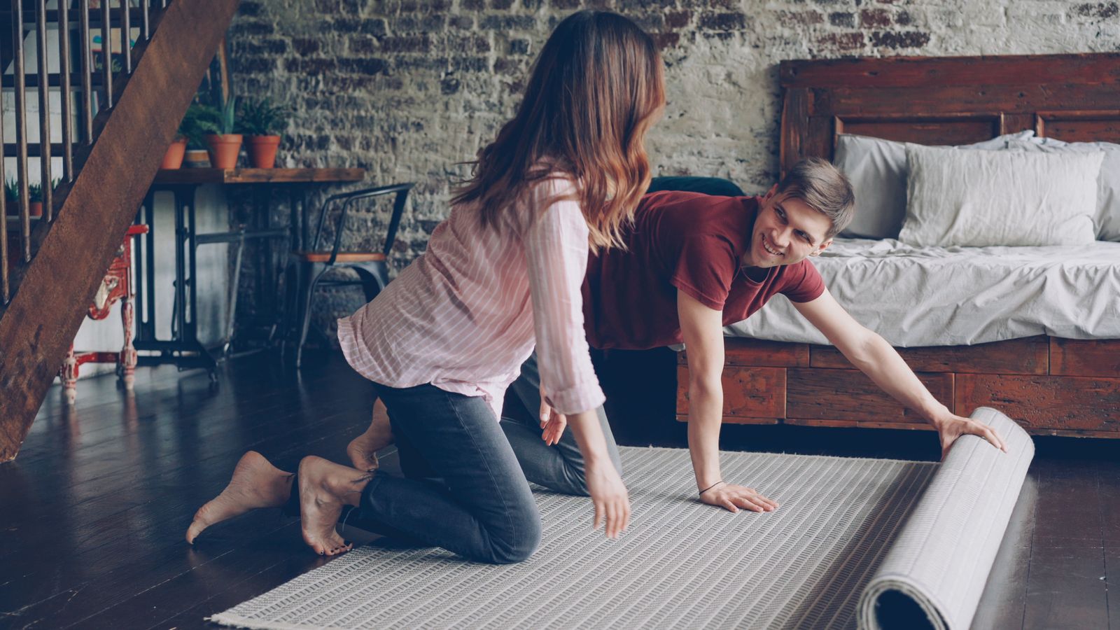 A couple unrolling a carpet in a stylish, industrial-themed bedroom with a brick wall.