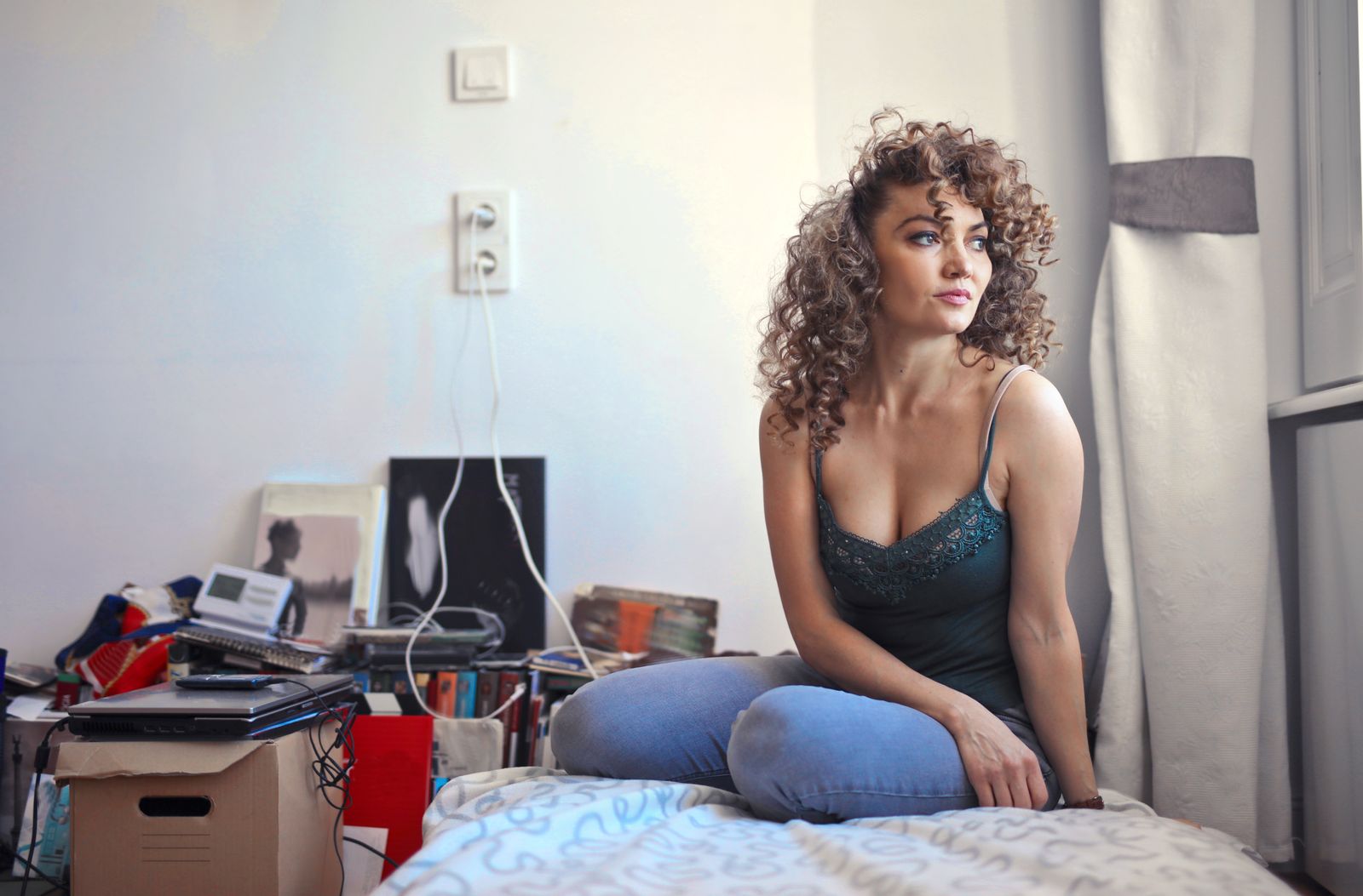 Curly-haired woman sitting pensively on a bed in a cluttered room, looking thoughtful.