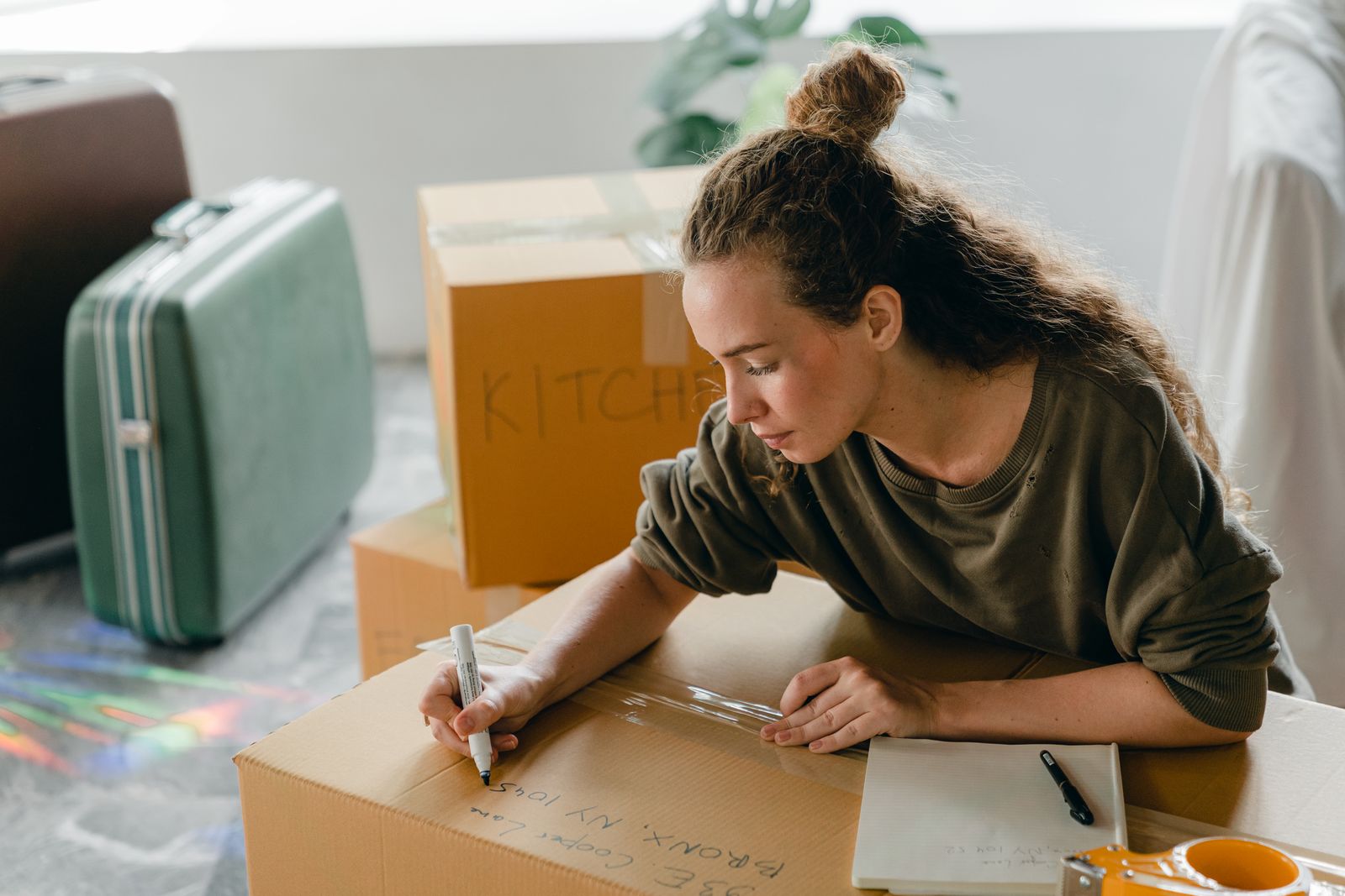 Top view of thoughtful lady in casual wear writing with marker on cardboard box while sitting near suitcases and pile of boxes in light living room