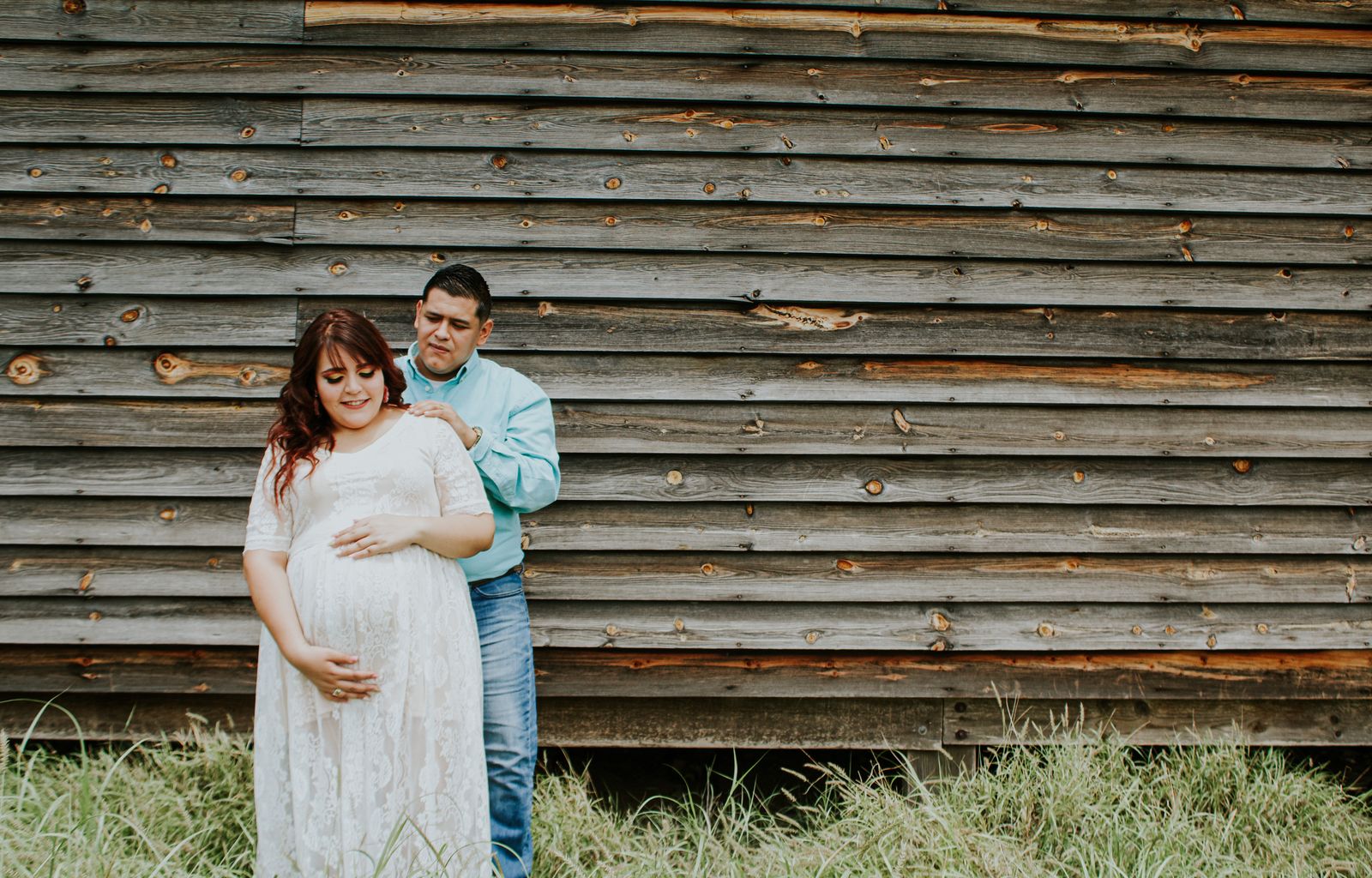 Young cheerful expectant female in elegant dress touching belly behind male partner on grass near old ribbed wall