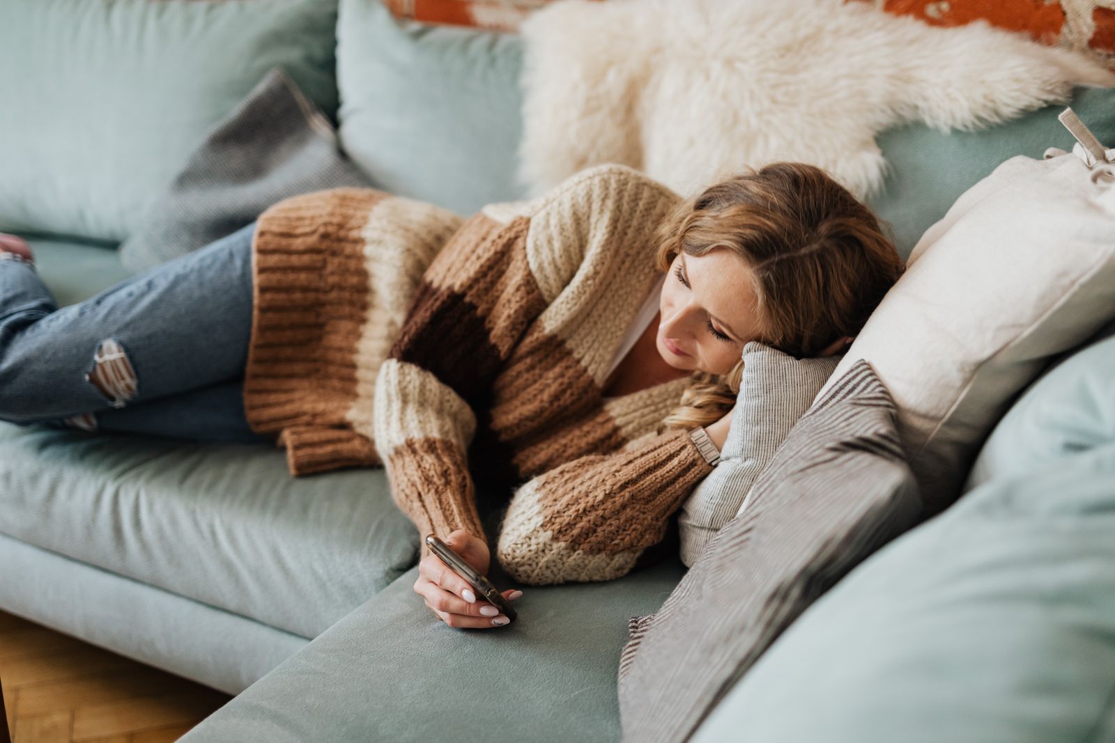 A woman in a cozy sweater relaxing on a sofa while using her phone indoors.
