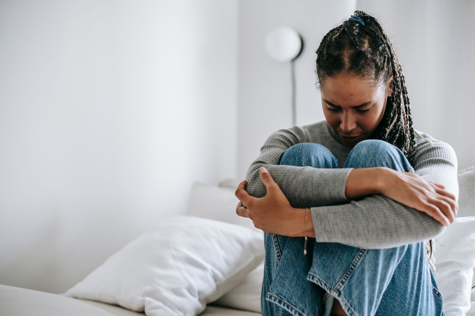 Disappointed ethnic female embracing legs while suffering from problem and sitting on white bed