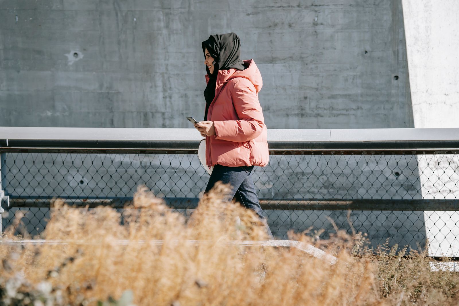 Side view of young Muslim female in casual wear and traditional headscarf walking along metal fens and concrete wall on street