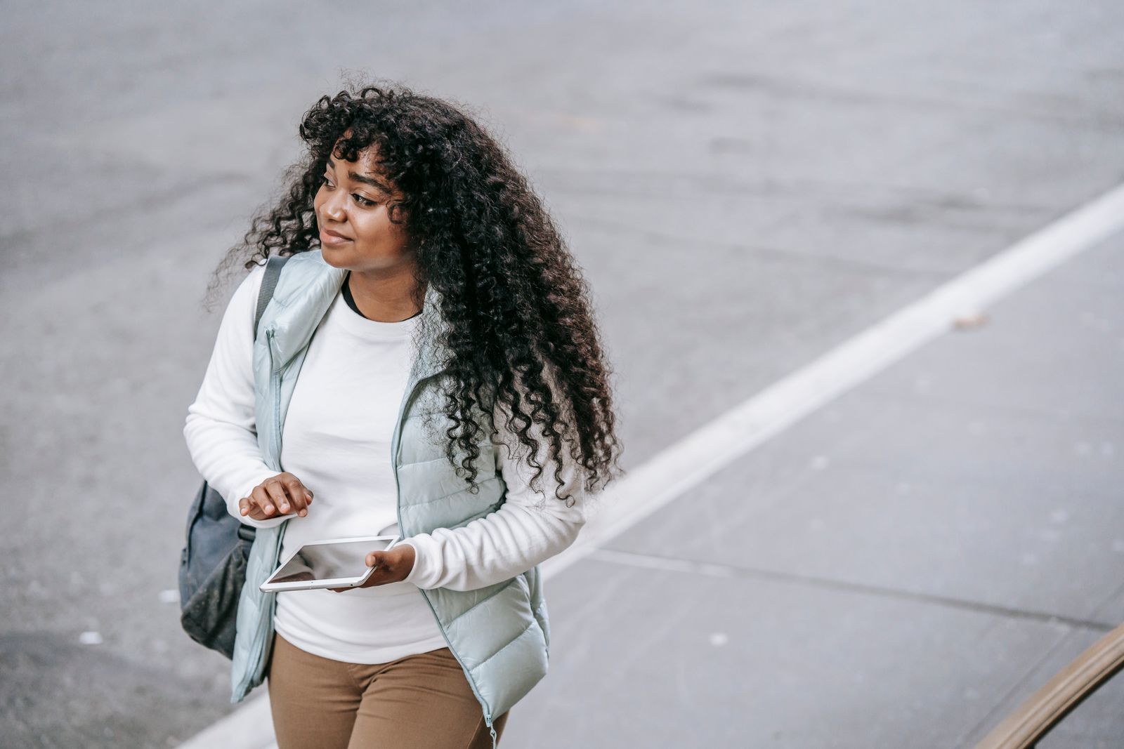 Young black lady wearing casual clothes and backpack while strolling on city street with tablet on walkway near road in daytime and looking away