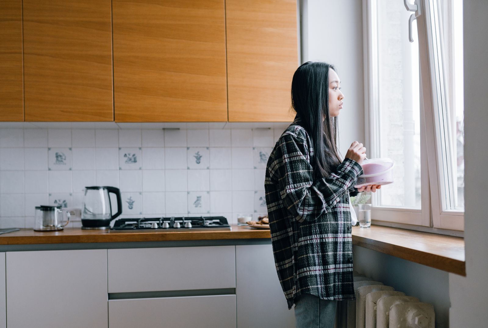 A woman stands in her kitchen, thoughtfully gazing out the window, holding a bowl.