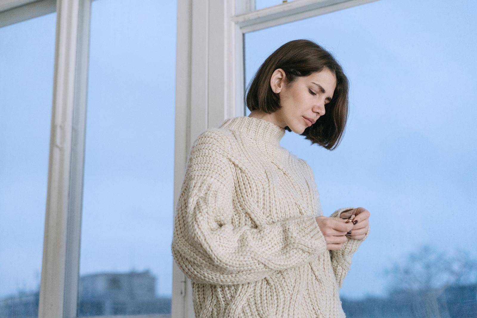 A woman in a cozy knit sweater stands contemplatively by a window.
