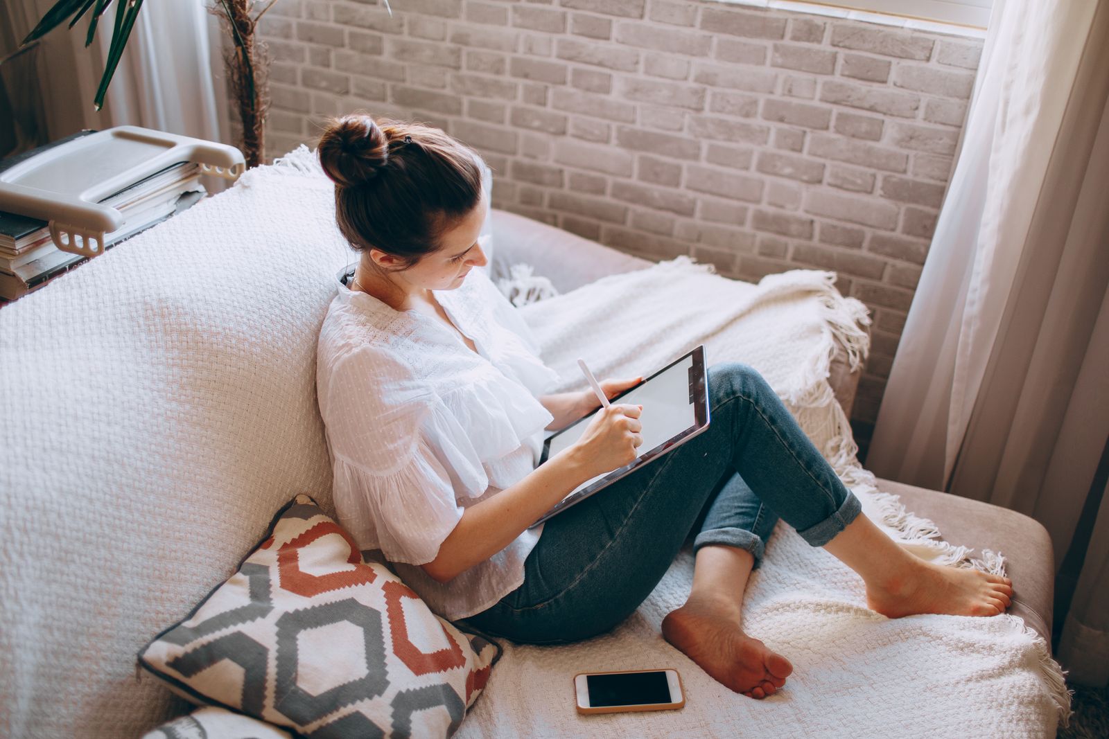 From above of young focused female in casual clothes sitting on sofa and drawing illustrations in tablet at home