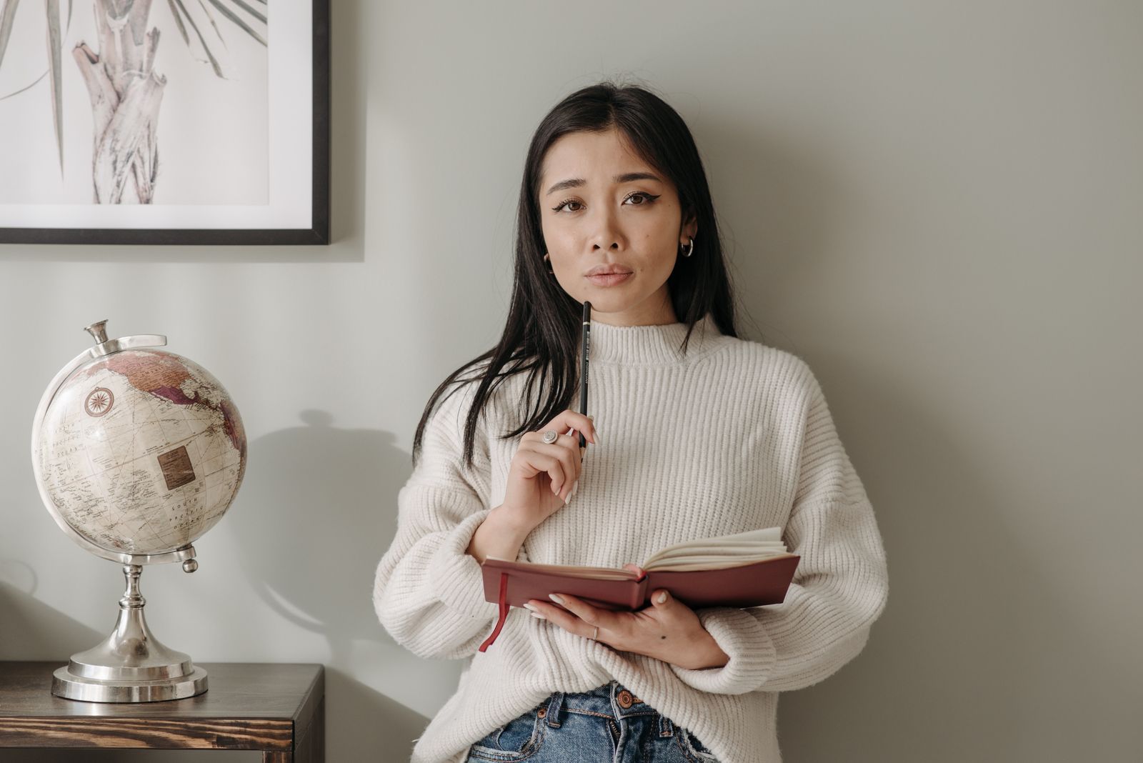 Asian woman in a cozy sweater holds a notebook and pen, standing by a globe inside a room.