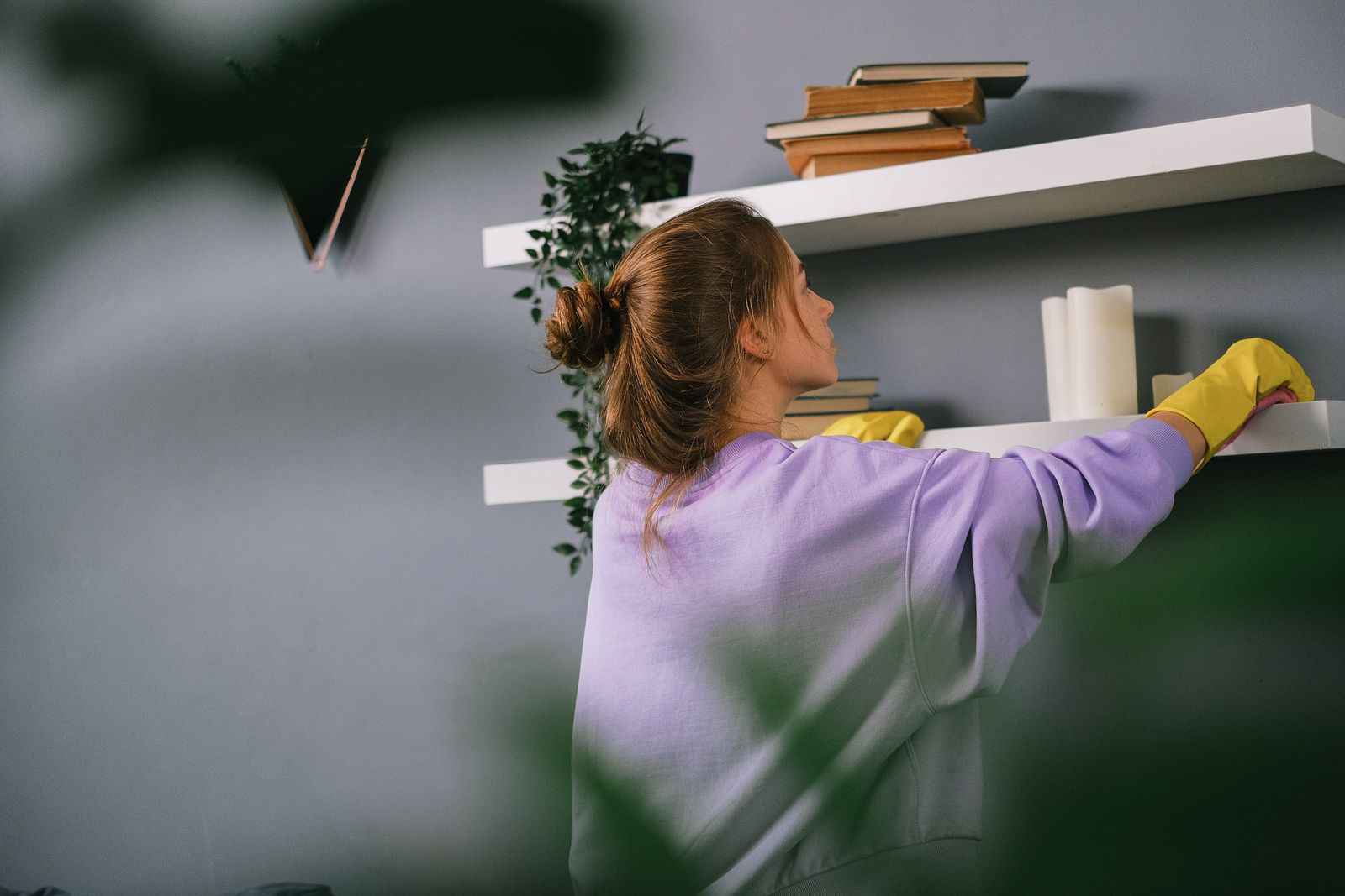 Side view of young woman in casual clothes and yellow gloves standing and wiping shelves with books and candles against gray wall in apartment