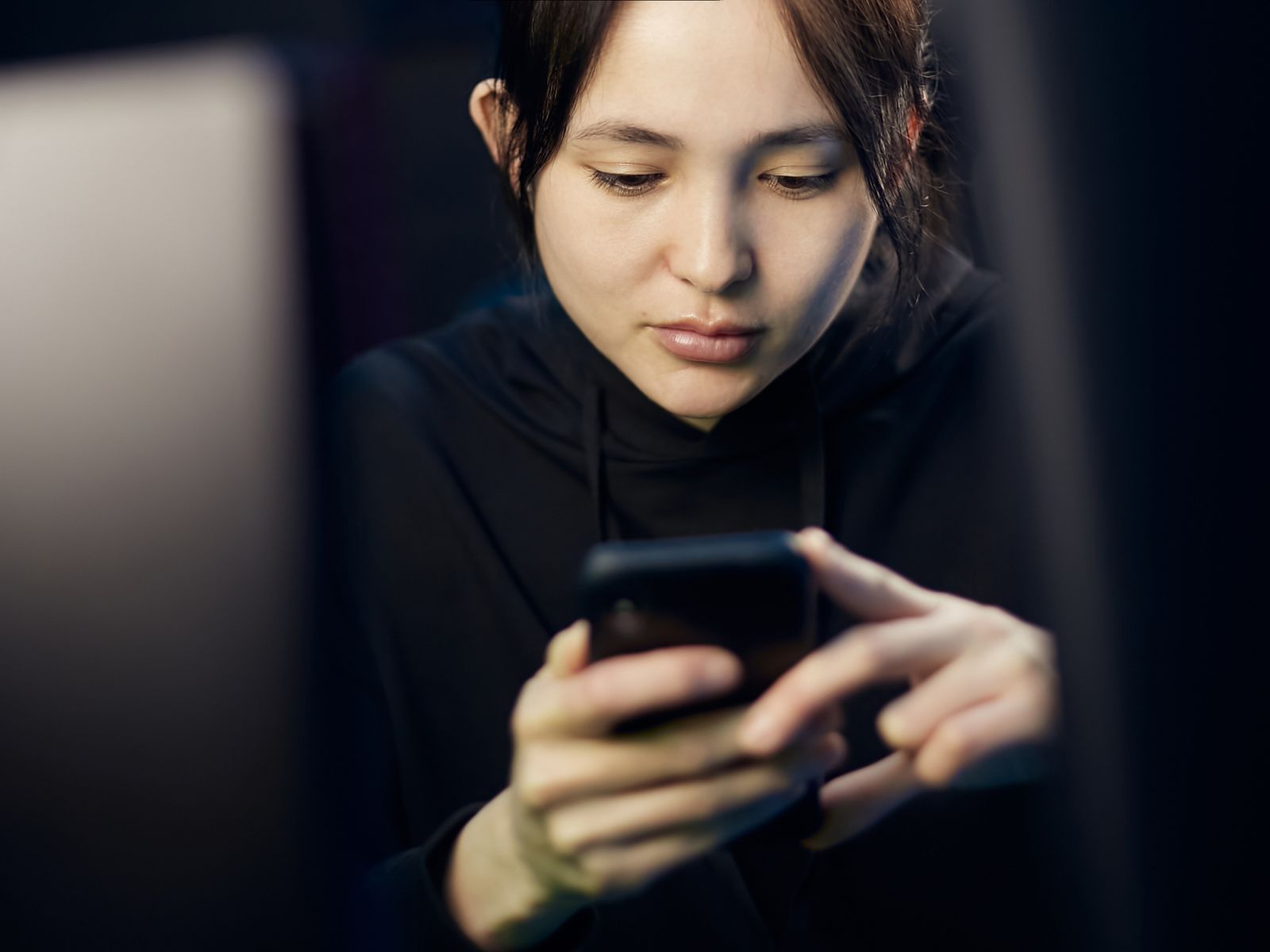 A woman intently looking at her smartphone while seated indoors, dim lighting.
