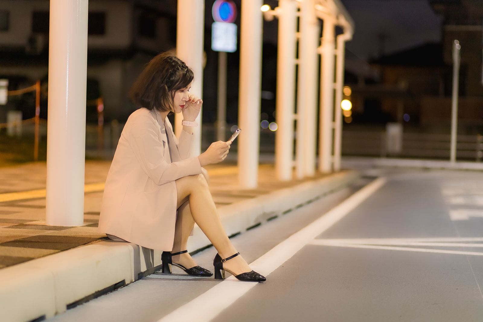 A woman sitting on a sidewalk using her smartphone at night under urban lights.