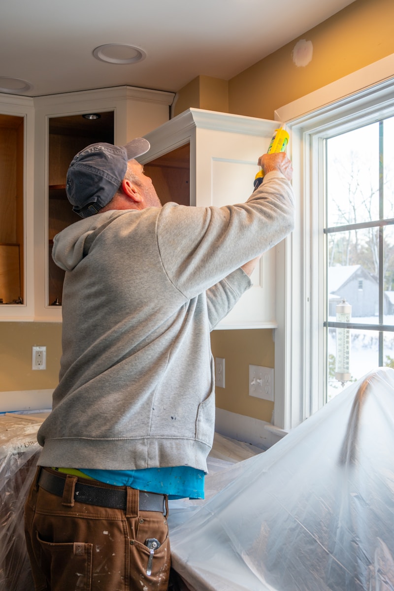 A person in a grey hoodie applying caulk to a window frame in a kitchen