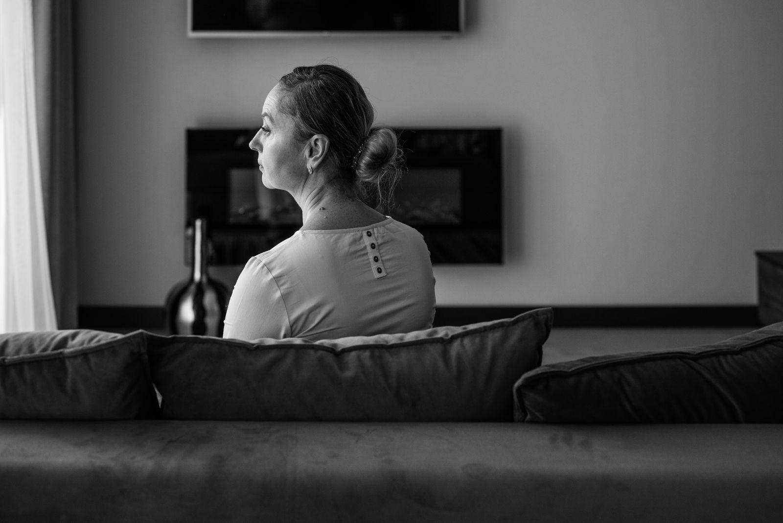 A senior woman sitting pensively on a couch, back view in a grayscale setting.