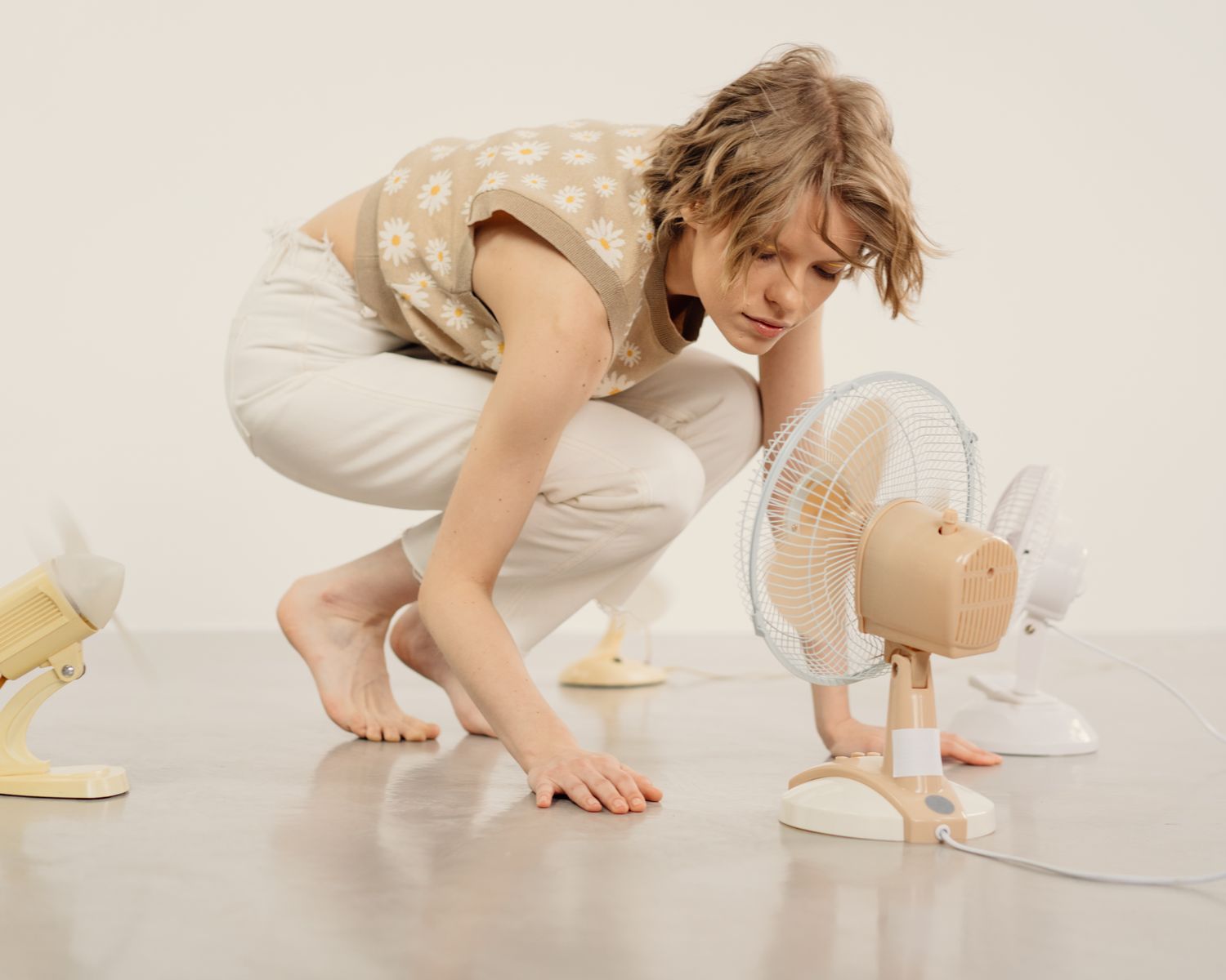 Young woman observing several table fans in minimalist indoor studio setting.