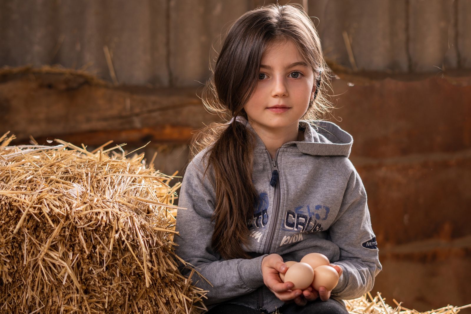A young girl with a ponytail and hoodie sits on hay, holding eggs inside a rustic barn setting.