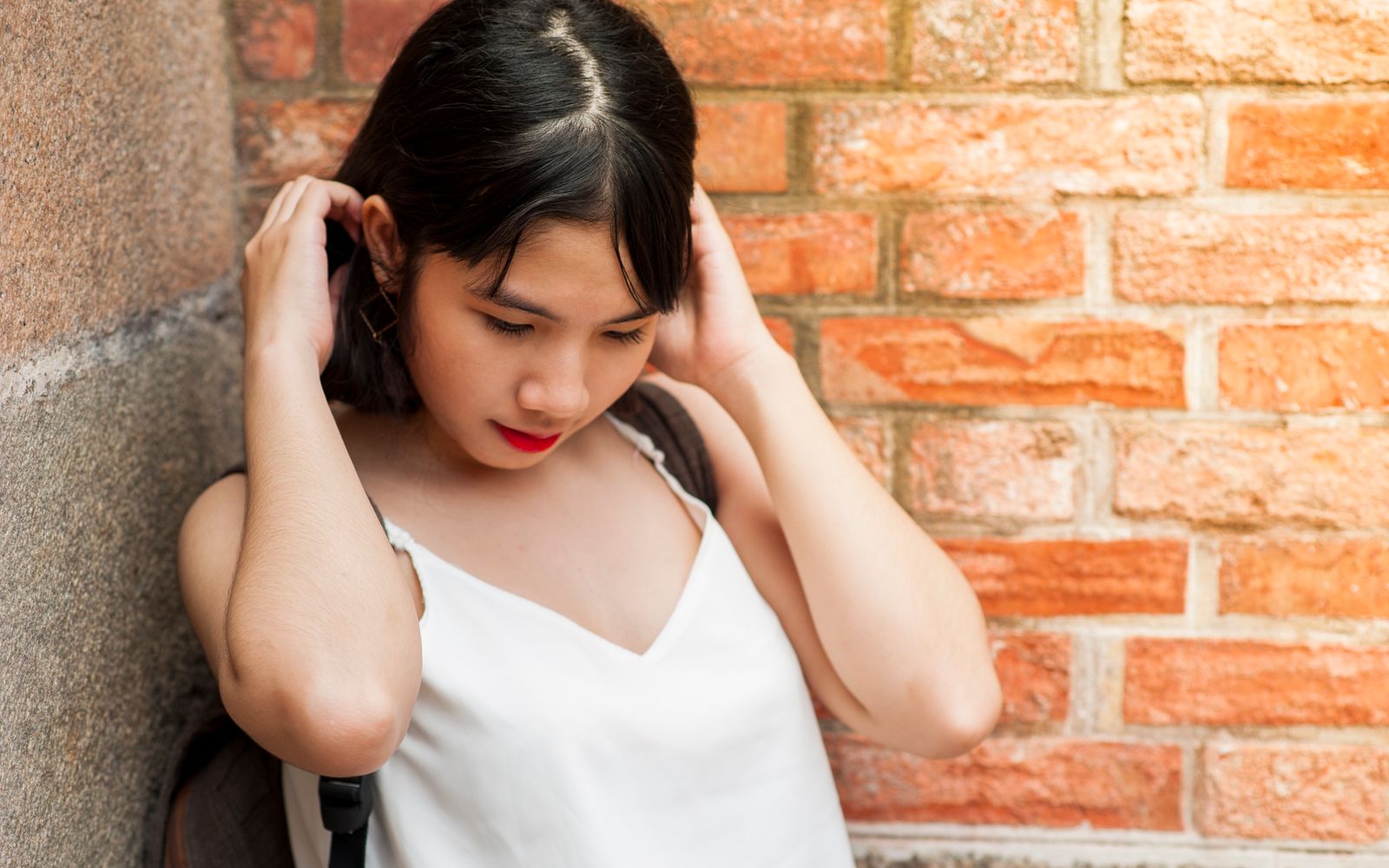 Stylish young woman in white top poses thoughtfully against a rustic brick wall.