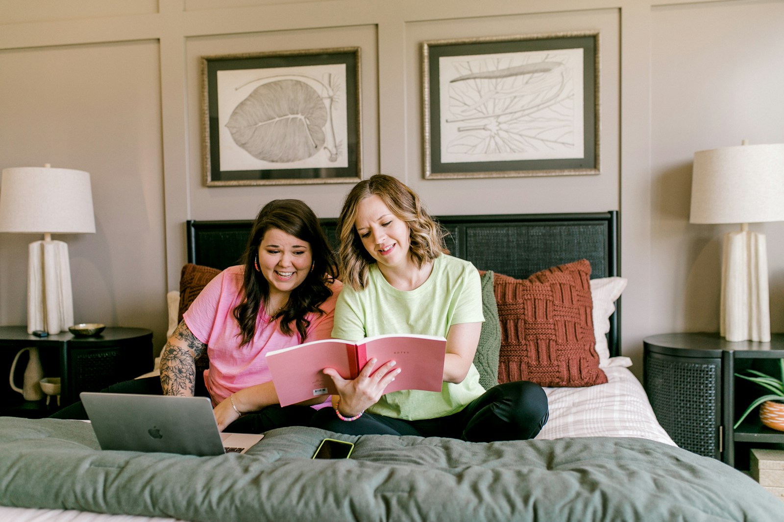 Two smiling women relaxing on bed with laptop and book.
