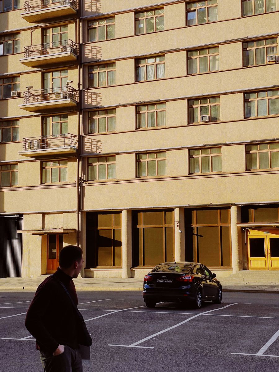 Man walking past a modern apartment building facade with parked car.