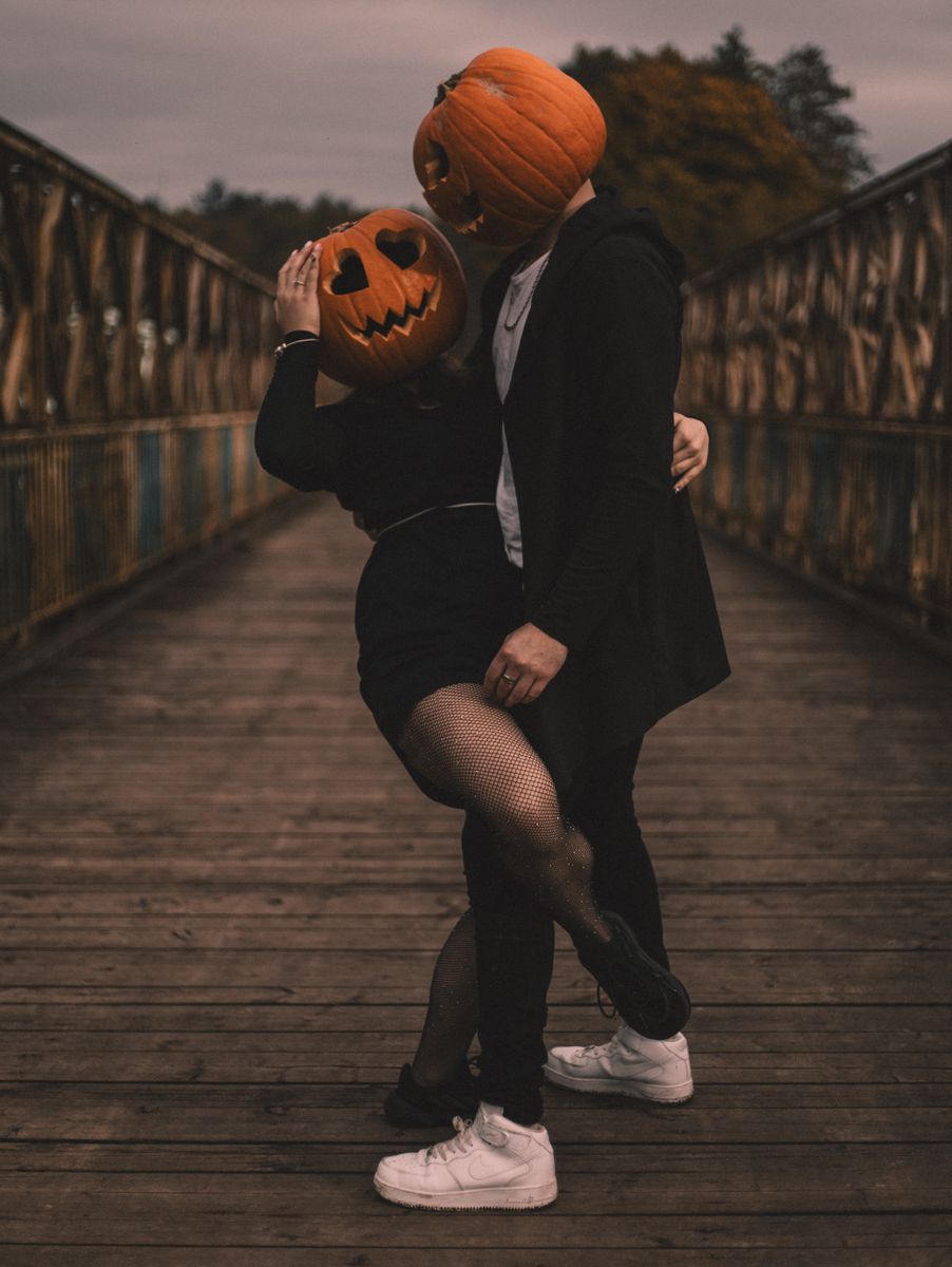 A couple wearing pumpkin heads embraces on a wooden bridge, capturing the eerie mood of Halloween.
