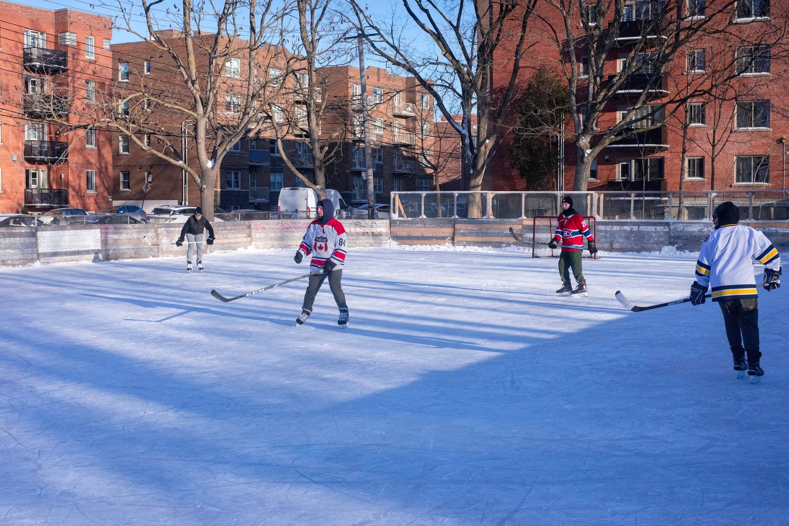 Adults playing ice hockey on an outdoor rink in a city during winter.
