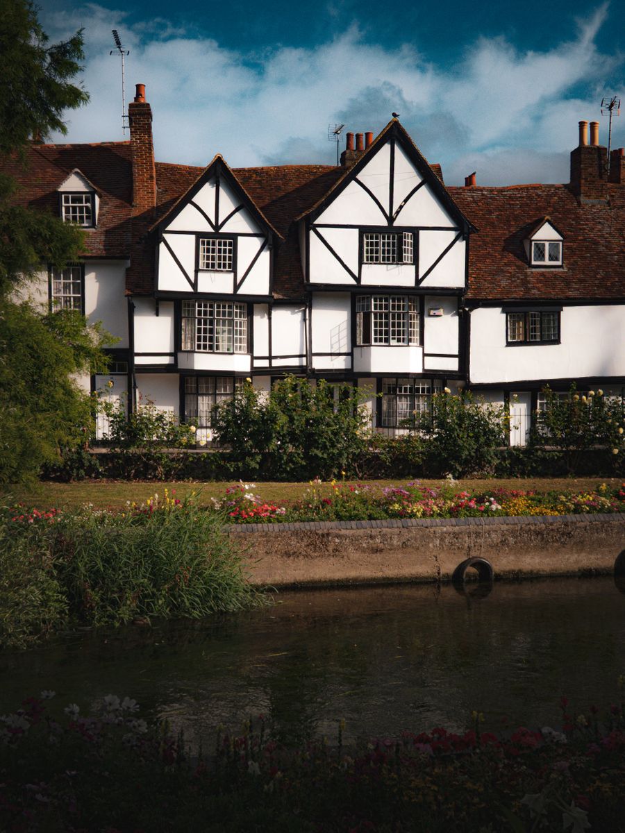A picturesque Tudor-style house with lush gardens by a tranquil river under a bright blue sky.