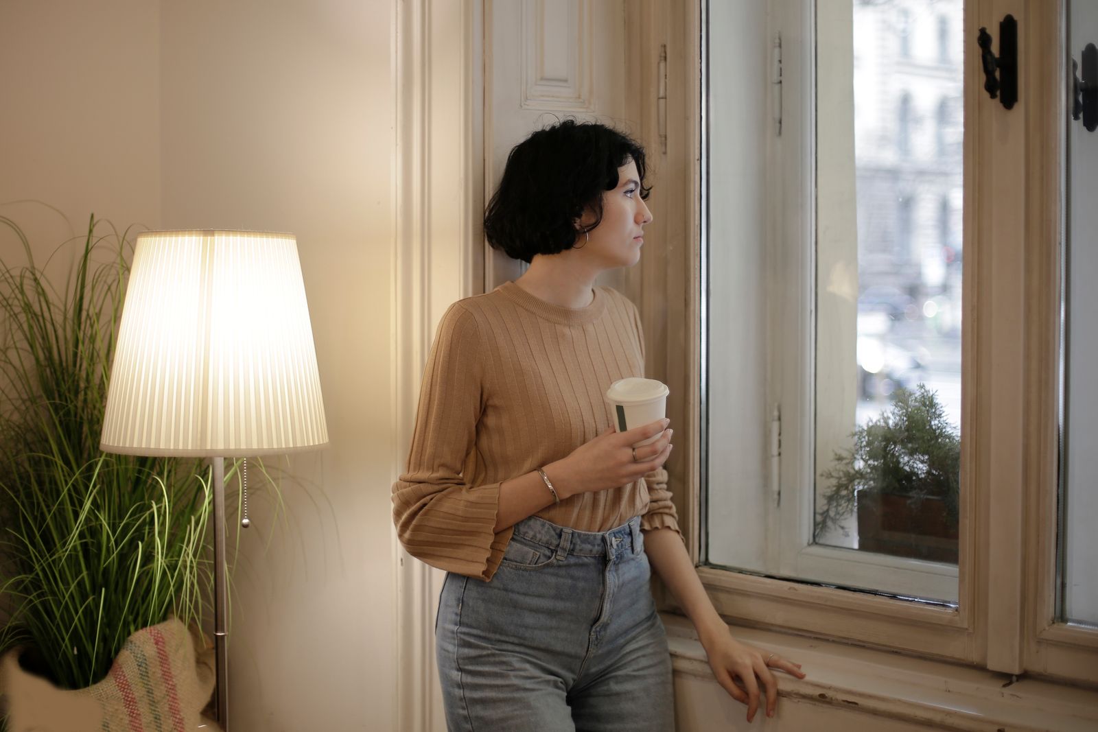 Young woman holding a coffee cup, gazes thoughtfully out the window in cozy room.