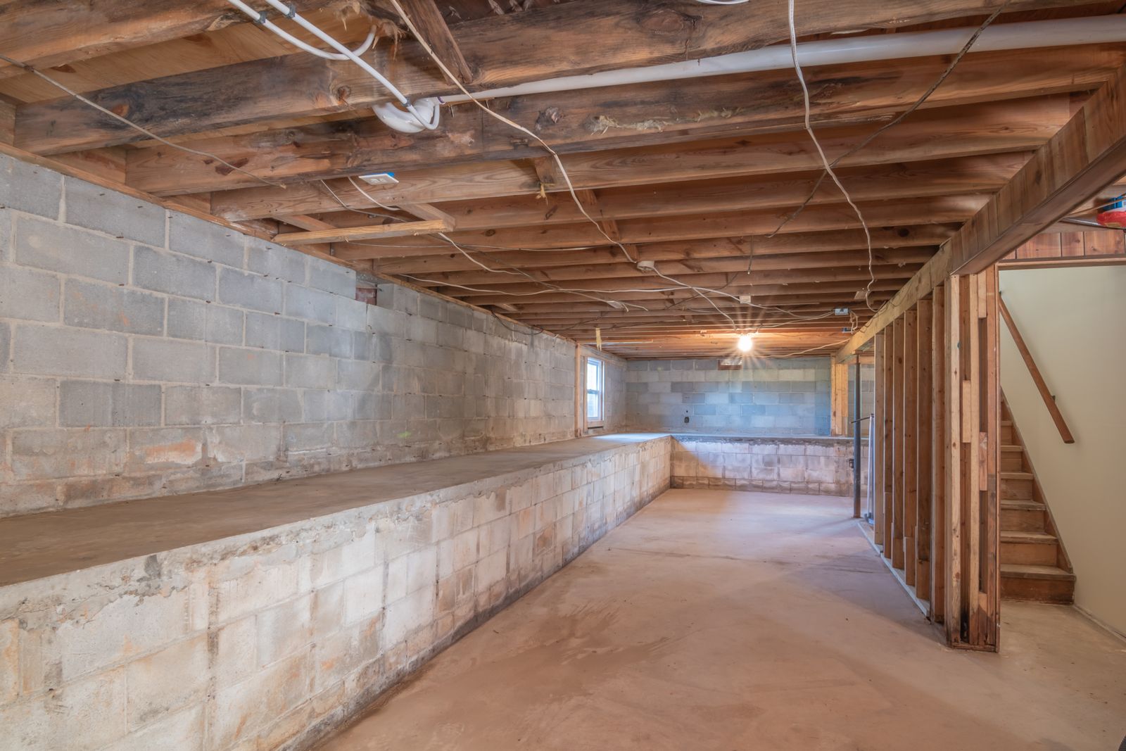 Empty basement with exposed wooden beams, concrete walls and stairs.