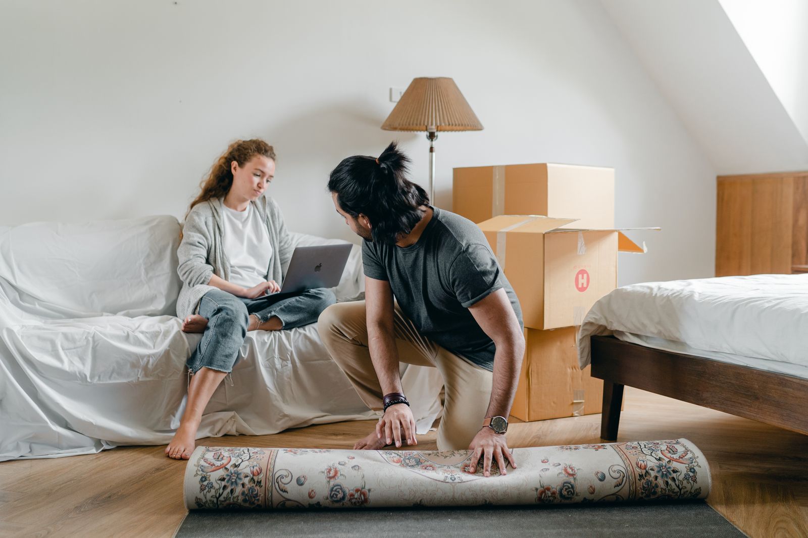 Pensive barefoot girlfriend surfing internet on netbook while sitting on comfortable couch and ethnic man in smart watch laying rug on floor after moving into new house