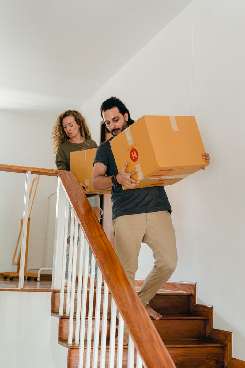 Full body of young woman and barefoot ethnic man in casual clothes carrying boxes on wooden stairs while moving into new home