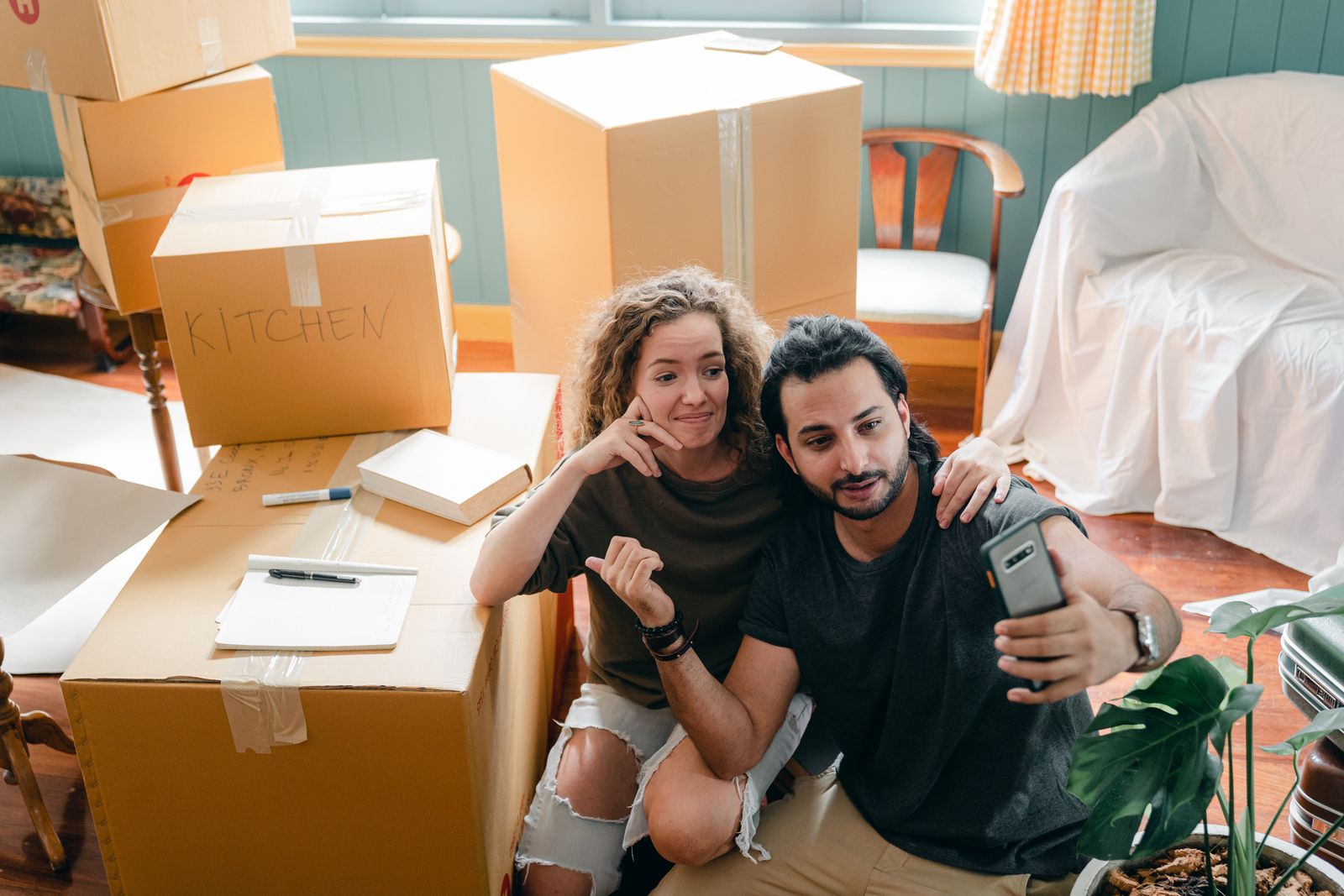 Top view of trendy woman embracing ethnic boyfriend in casual wear while taking selfie on cellphone and sitting surrounded by heap of cardboard boxes near sofa covered with bed sheet