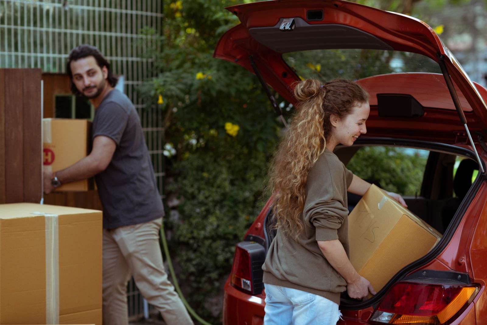 Cheerful young woman with curly hair taking cardboard package out from trunk of automobile while bearded ethnic man carrying box into new house in suburb or countryside area