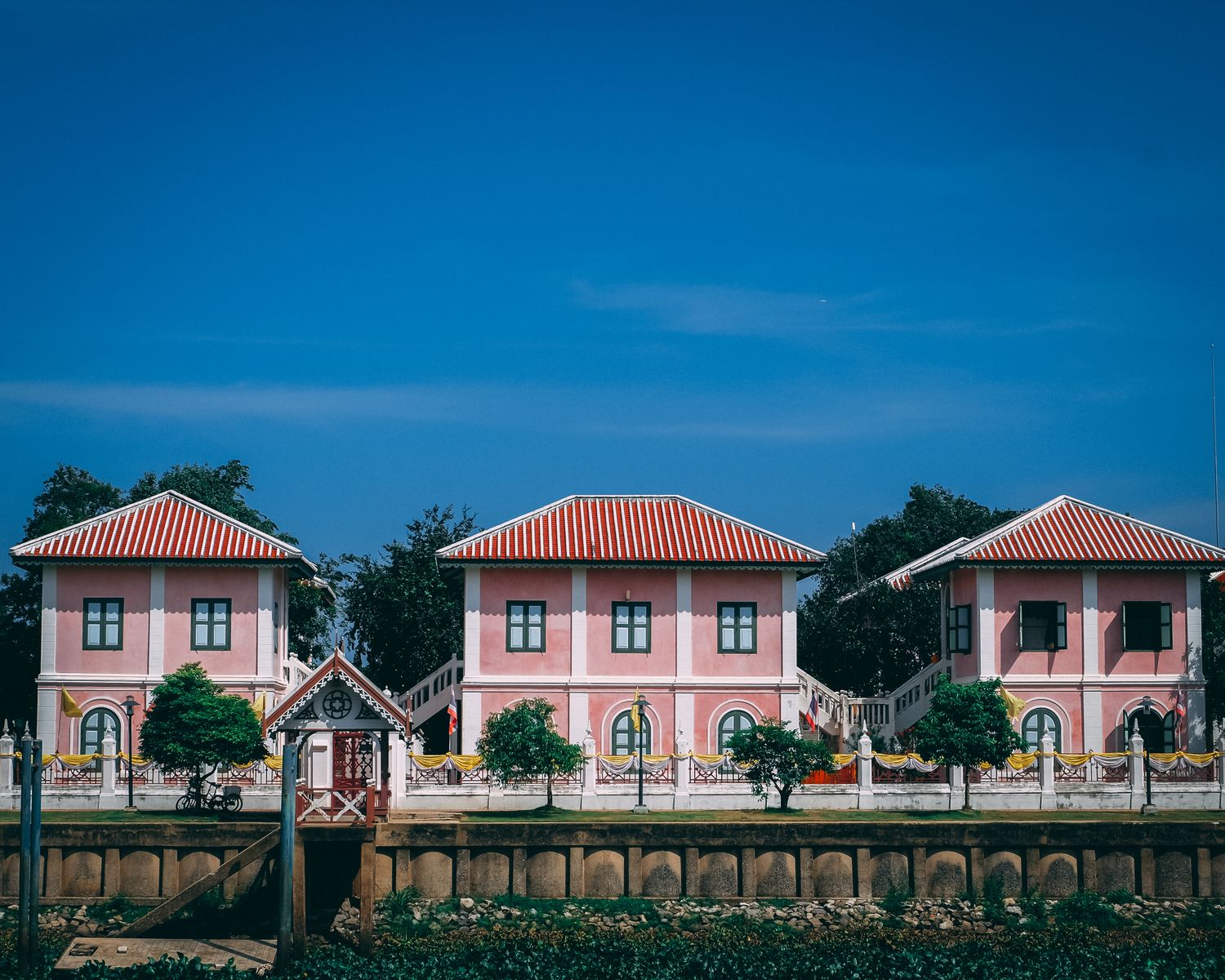Similar aged building facades with ribbed roofs near fence and trees under colorful cloudless sky