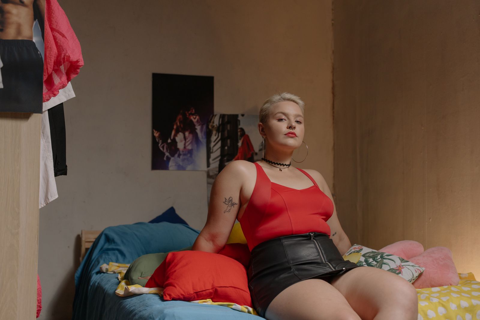 A confident woman with short blond hair poses on a bed in a stylish black skirt and red top, showcasing tattoos and fashion accessories.