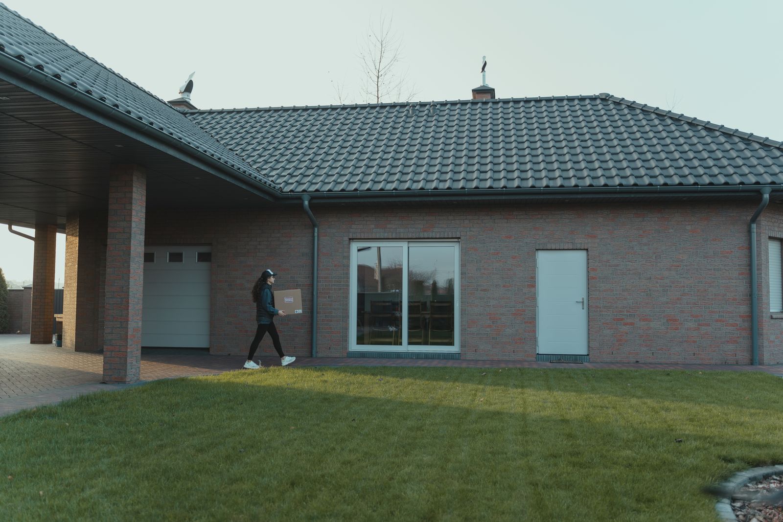 Person delivering package to a modern brick house with a tile roof and green lawn.