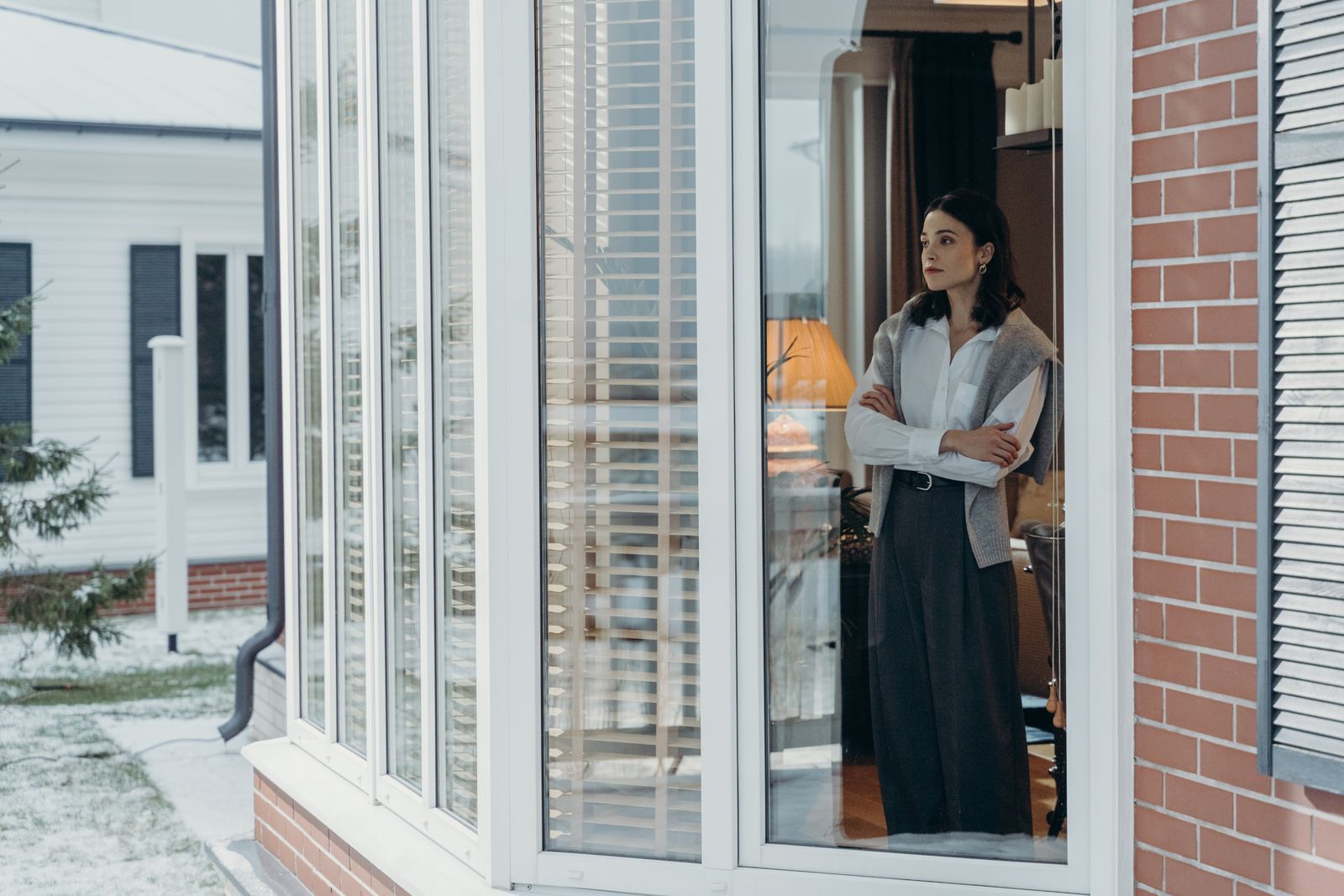 Contemplative woman stands with arms crossed, gazing out a window. Thoughtful and serene.