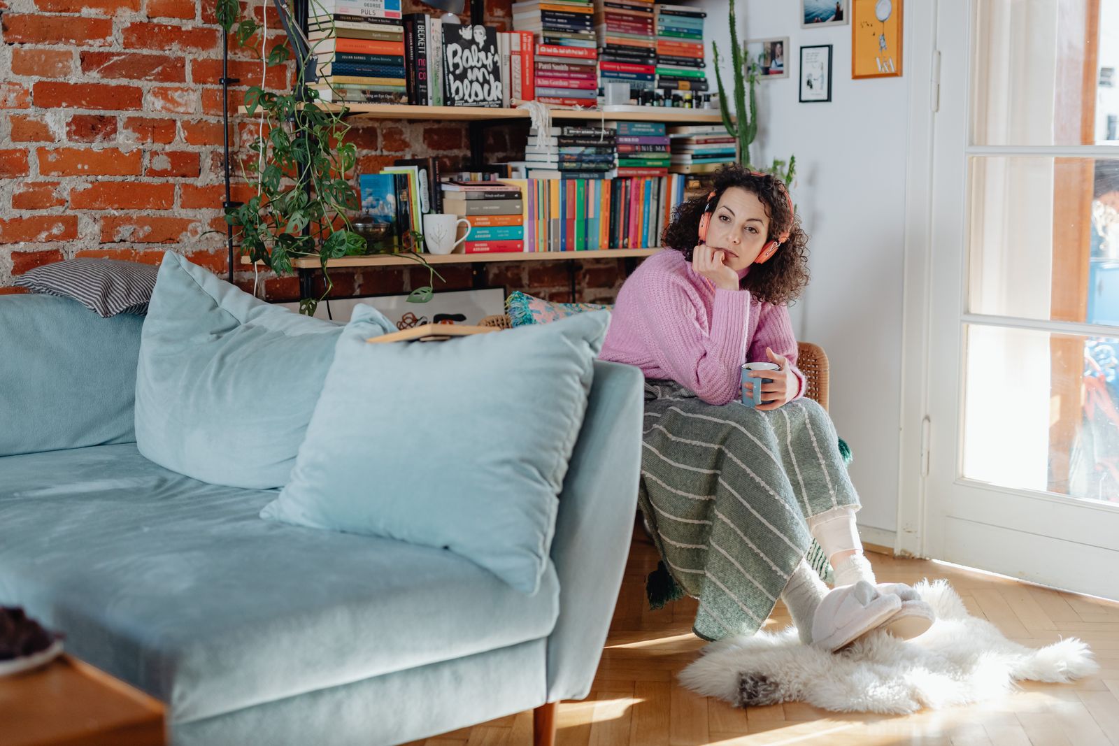 Woman sitting in cozy living room with headphones, lost in thought, cradling a warm cup.