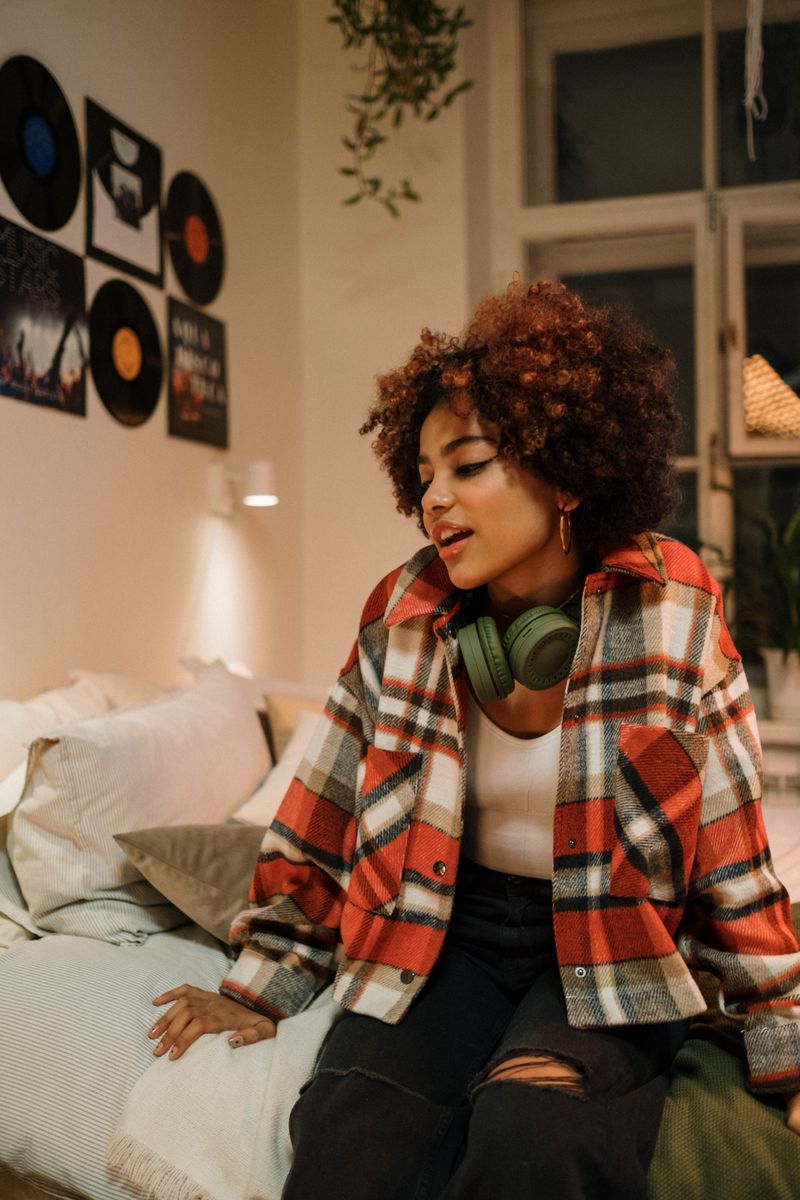 Young woman in plaid shirt relaxing in bedroom with headphones, cozy decor around.