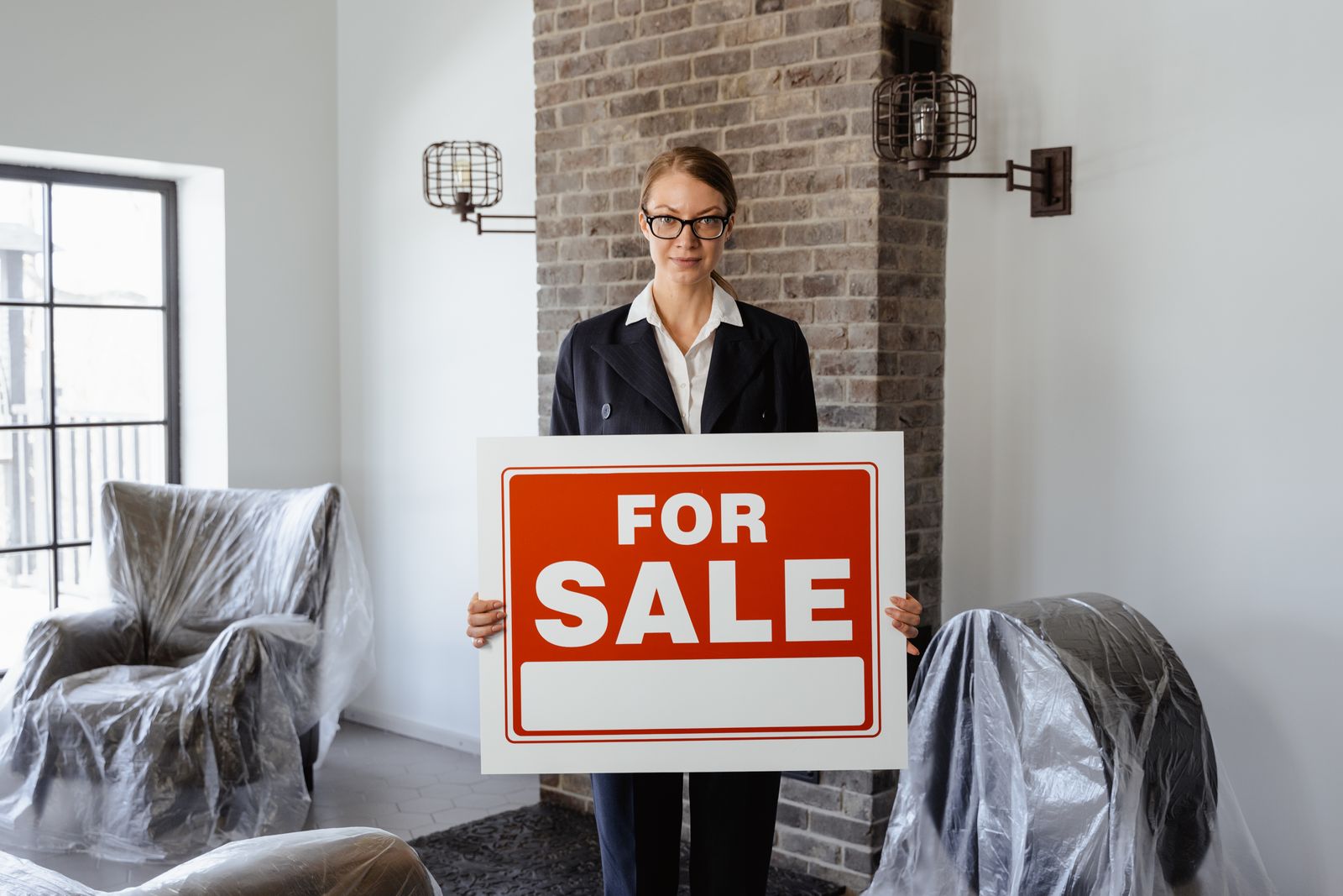 Real estate agent holding a 'For Sale' sign in a furnished living room.