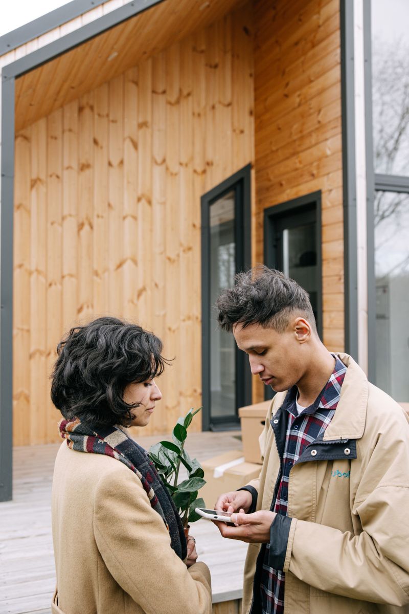 Young couple standing outside their new wooden home holding a plant and smartphone.