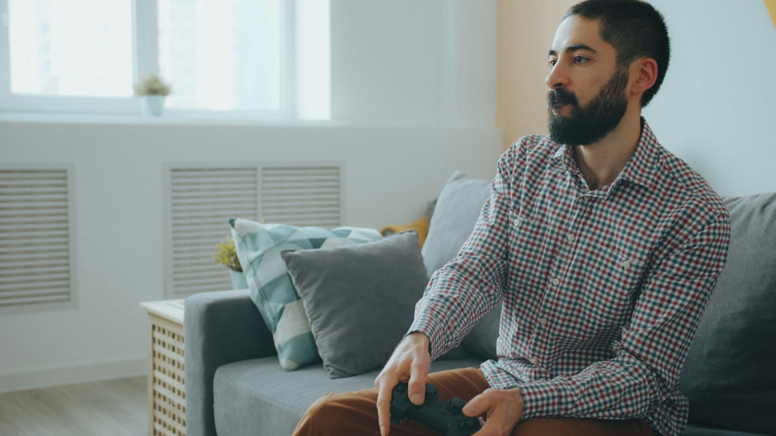 A man with a beard in a plaid shirt playing video games on a sofa
