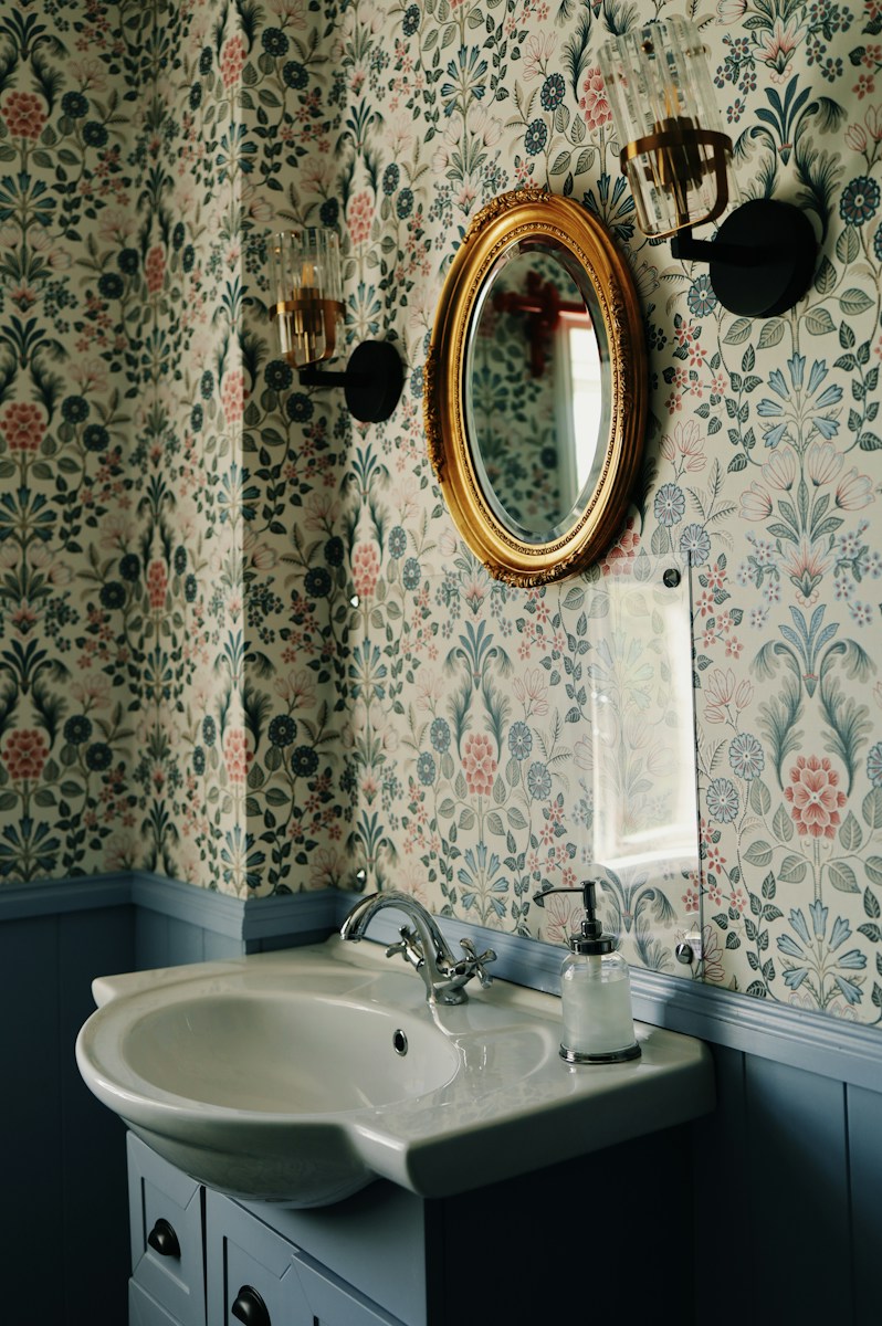 A bathroom with floral wallpaper and gold mirror