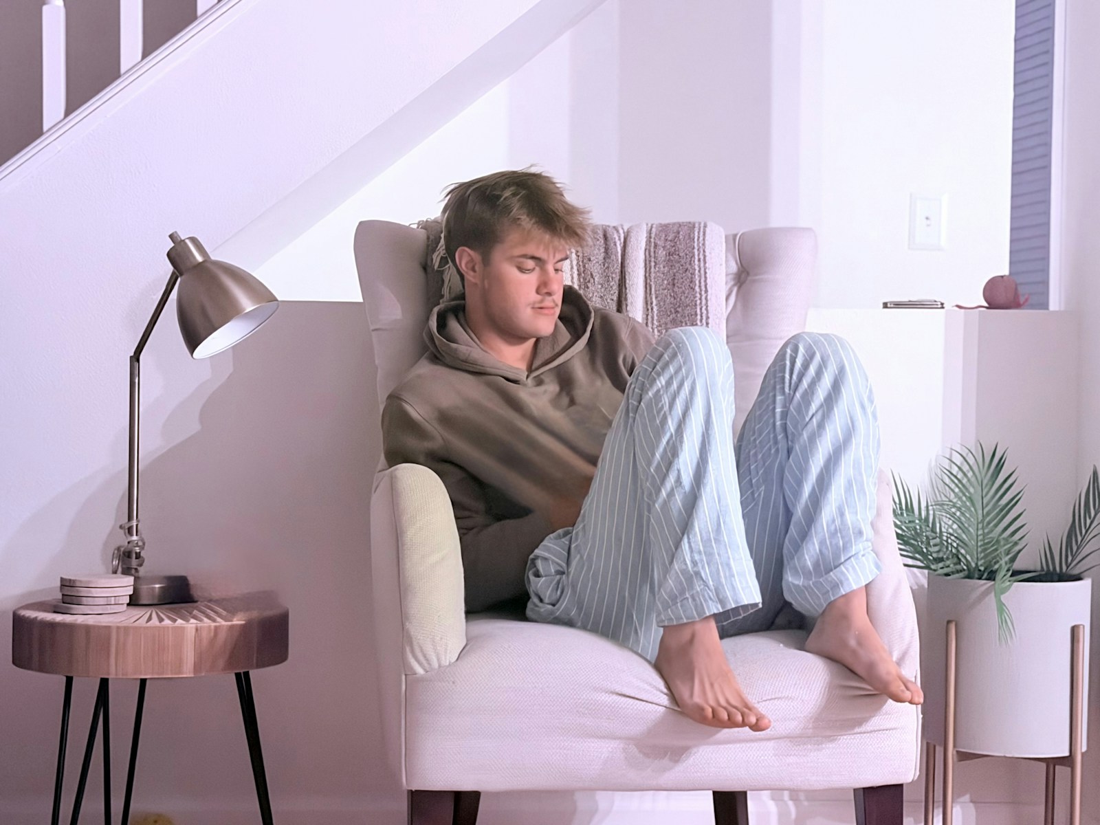 A young man in a hoodie and striped pajama pants curled in an armchair