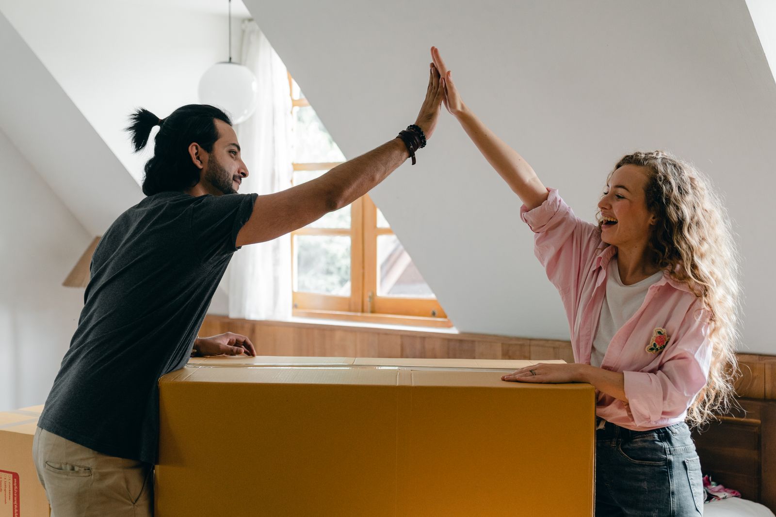 Side view of cheerful young ethnic bearded man with ponytail and woman with curly hair giving high five and looking at each other standing with long cardboard box in attic bedroom of new house