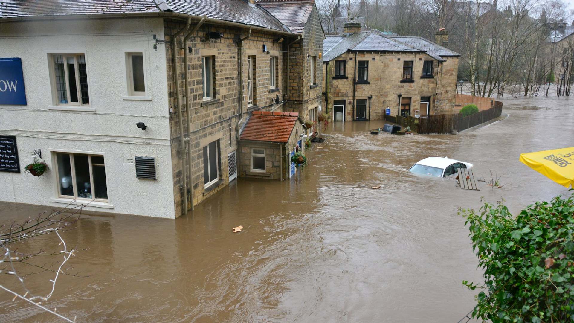 Floodwaters surrounding stone buildings and a partially submerged white car in a town. Photo by Chris Gallagher on Unsplash.